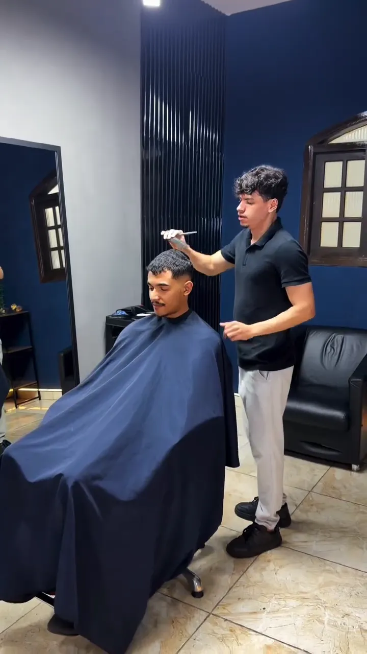 Interior do studio com parede azul marinho: o barbeiro Kauê penteia um cliente sentado na cadeira com capa azul